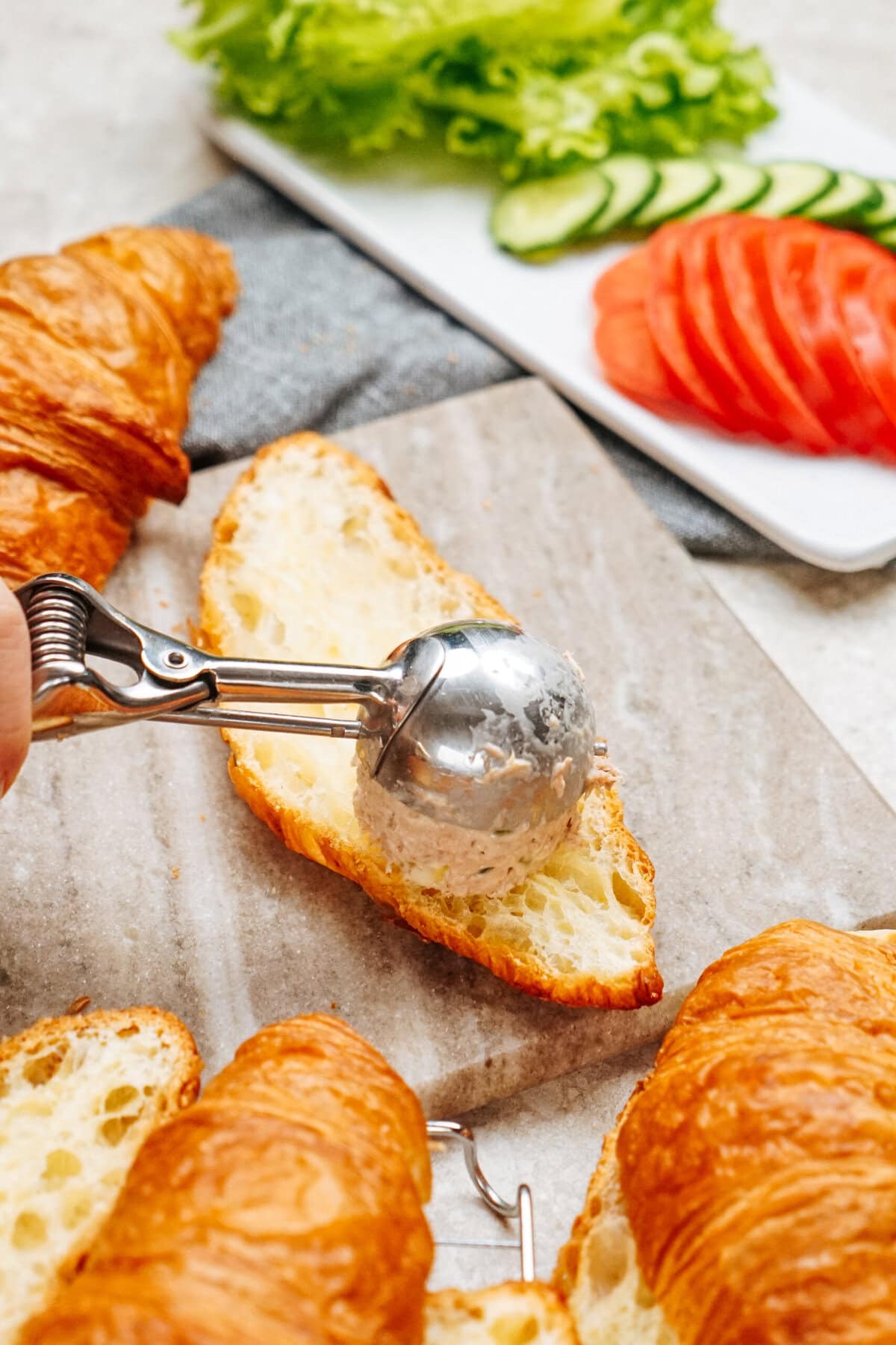 A scoop of tuna salad spread is placed on a sliced croissant. In the background, there are sliced cucumbers, tomatoes, and crisp lettuce on a plate.