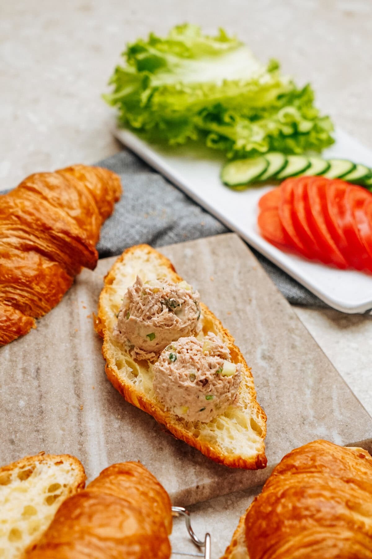 A slice of croissant serves as the elegant base for a delightful tuna salad sandwich, elevated by two generous scoops. In the background, a fresh plate of lettuce, cucumber slices, and tomato slices completes the scene.