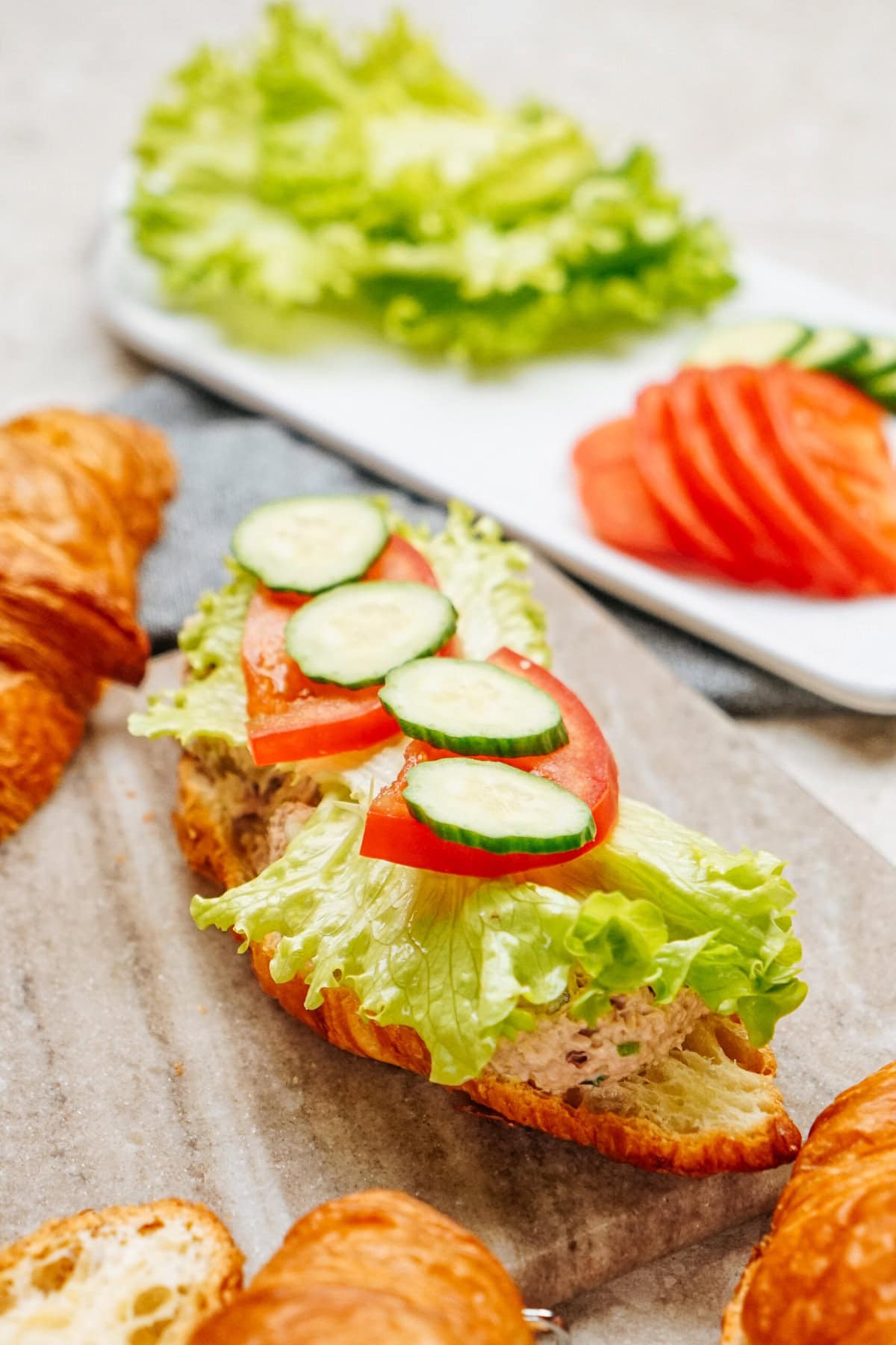 A croissant sandwich with lettuce, tomato, cucumber slices, and a delicious tuna salad filling sits on a board. More lettuce and tomato slices accompany the tuna salad sandwich on a white plate in the background.