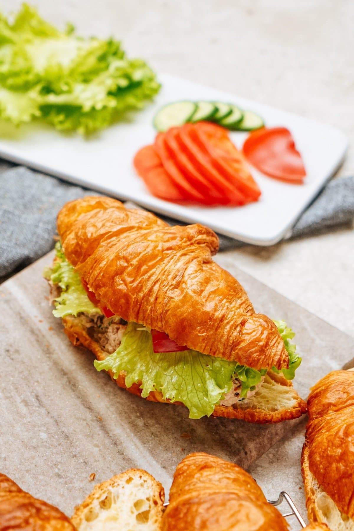 A croissant tuna salad sandwich with lettuce, tomato, and other fillings sits on parchment paper. Sliced tomatoes and cucumbers, and fresh lettuce leaves add a vibrant touch in the background.