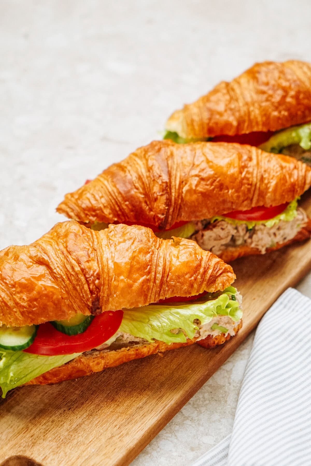 Three croissant sandwiches featuring a delightful tuna salad filling are artfully arranged on a wooden board, each accompanied by fresh lettuce, ripe tomato, and crisp cucumber.