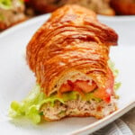 A croissant sandwich with lettuce, tomato, and a creamy tuna salad filling sits invitingly on a white plate.