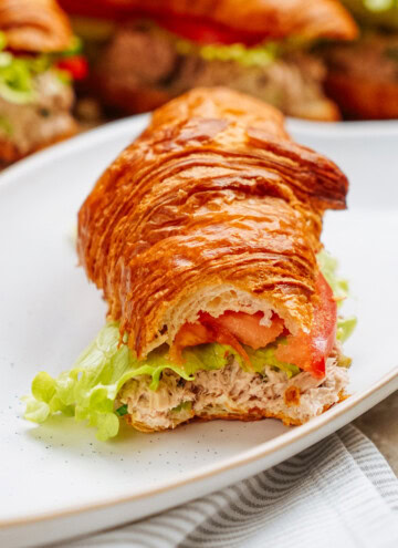 A croissant sandwich with lettuce, tomato, and a creamy tuna salad filling sits invitingly on a white plate.