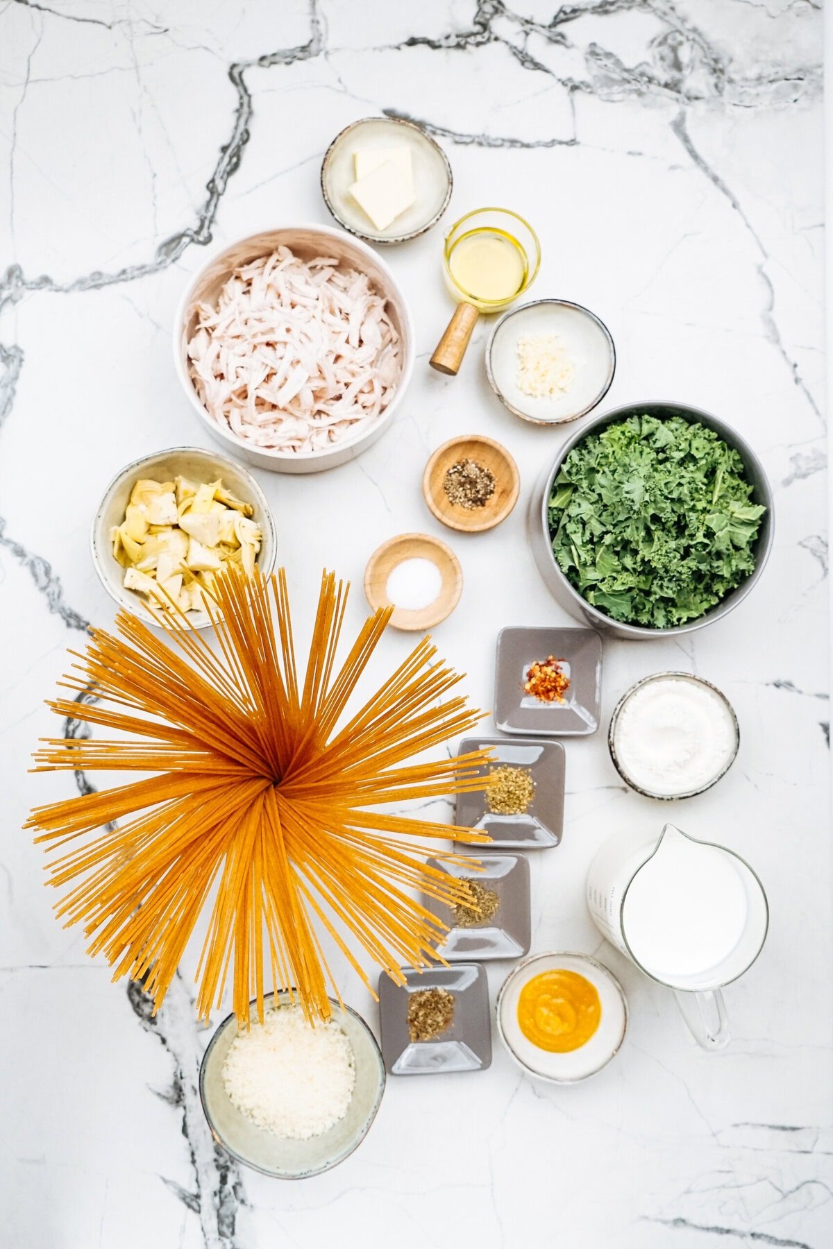 Top view of various ingredients arranged on a marble surface, showcasing chicken kale pasta essentials: uncooked spaghetti, shredded chicken, fresh kale, spices, cheese, and liquids in bowls and small dishes.