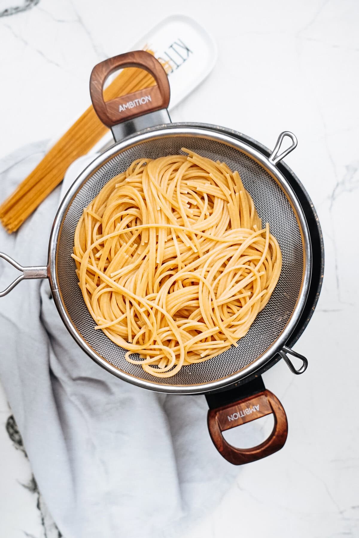 A colander with cooked spaghetti and tender chicken rests on a marble countertop, surrounded by uncooked pasta, fresh kale leaves, and a light gray cloth.