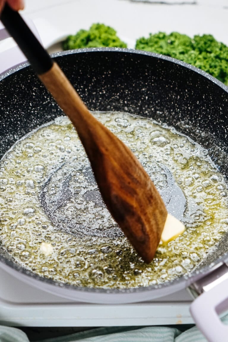 A wooden spatula stirs butter melting in a frying pan, setting the stage for a delicious chicken and broccoli dish. Kale peeks out in the background, promising a vibrant addition to the meal.