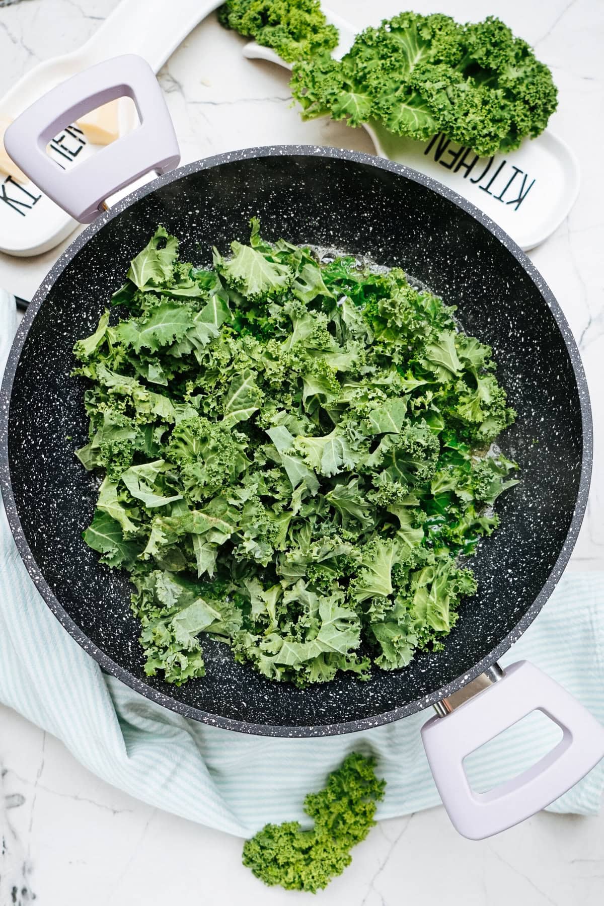 Fresh kale leaves in a frying pan on a marble surface, ready to be transformed into a delightful chicken and kale dish.