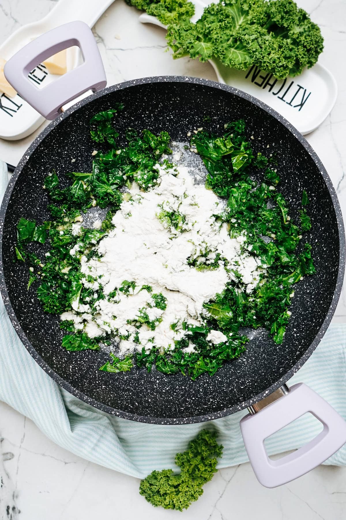 Chopped kale and flour sizzle in a large frying pan, while chicken adds a savory depth. In the background, extra kale and a kitchen-themed tray complete the culinary scene.