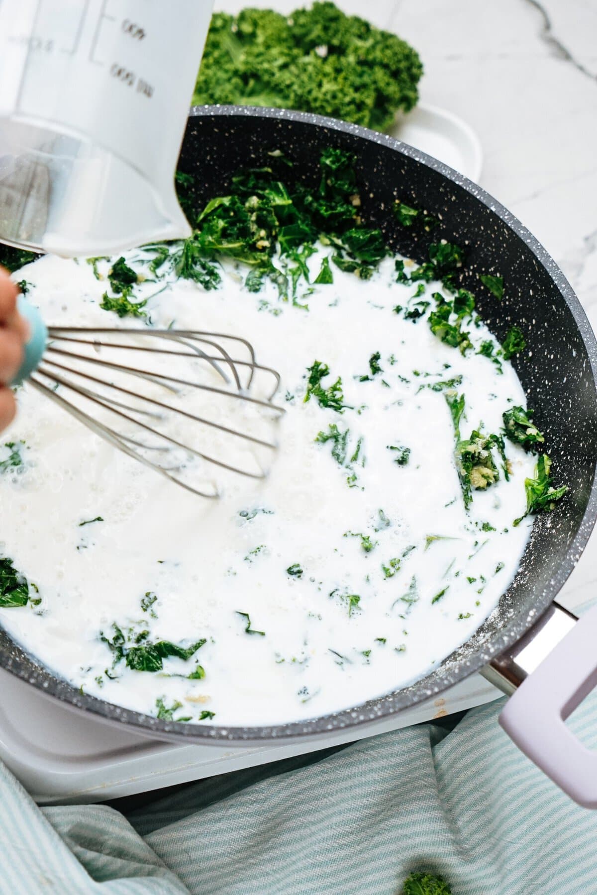 Whisking a creamy chicken mixture with chopped kale in a non-stick pan, next to a glass measuring cup.