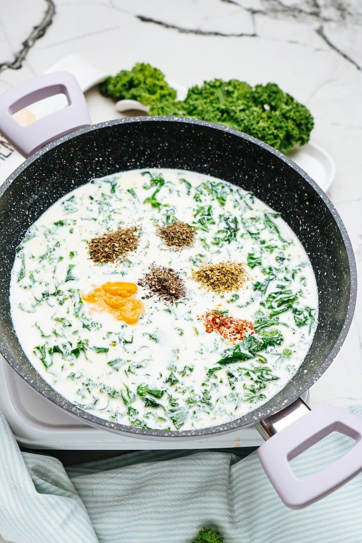 A skillet on a stove simmers a creamy chicken pasta mixture, topped with chopped greens and assorted spices. A fresh bunch of kale sits in the background, ready to enhance the dish with its vibrant flavor.