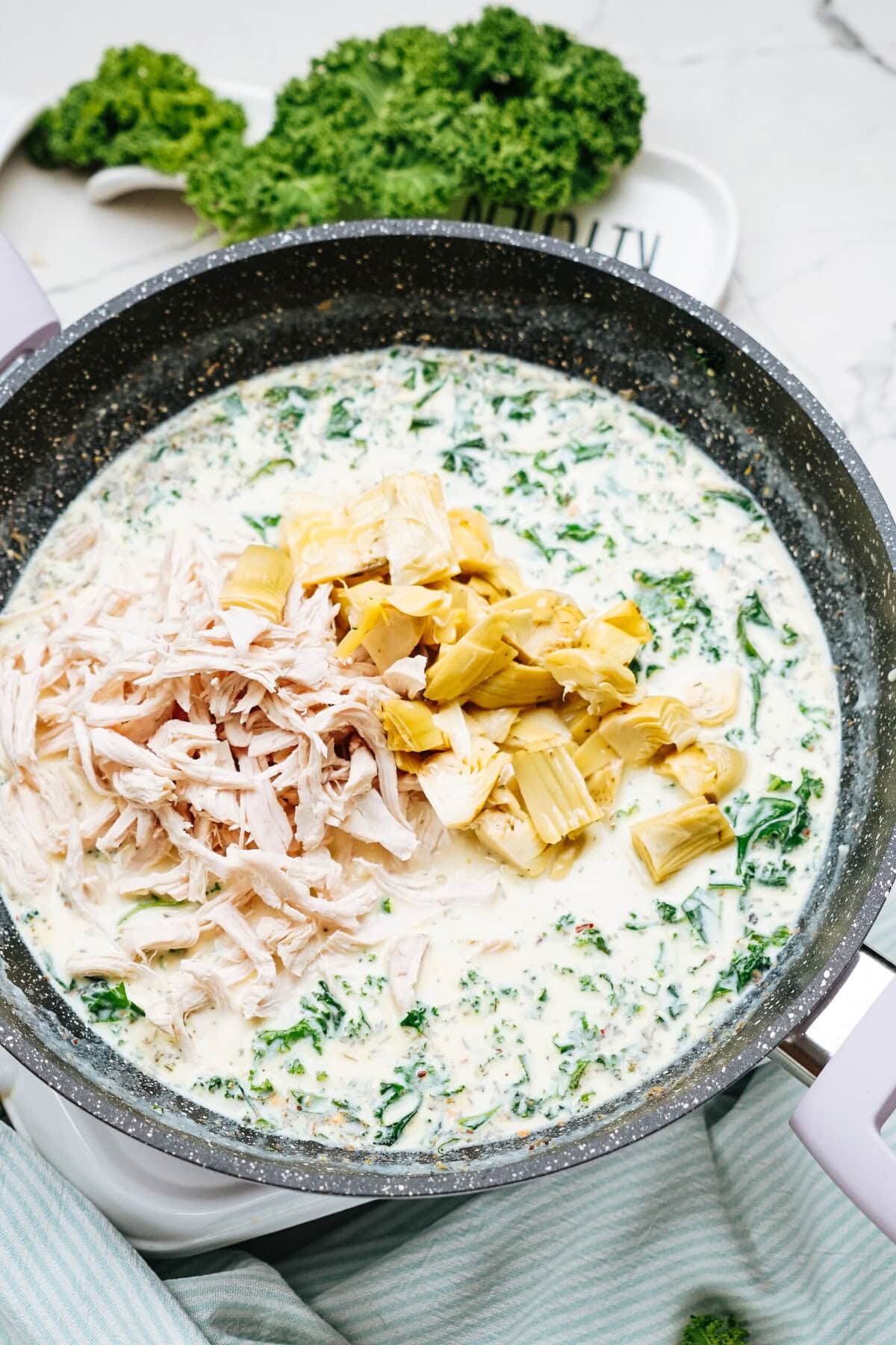 A pan filled with a creamy mixture containing shredded chicken, artichoke hearts, and chopped kale.