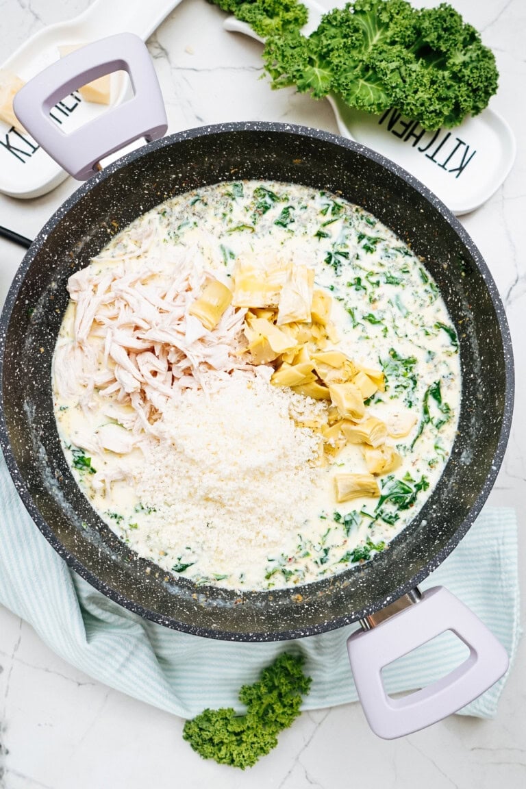 A black pot on a stove brims with creamy chicken kale pasta, topped with artichoke hearts and grated cheese, surrounded by kitchen utensils.