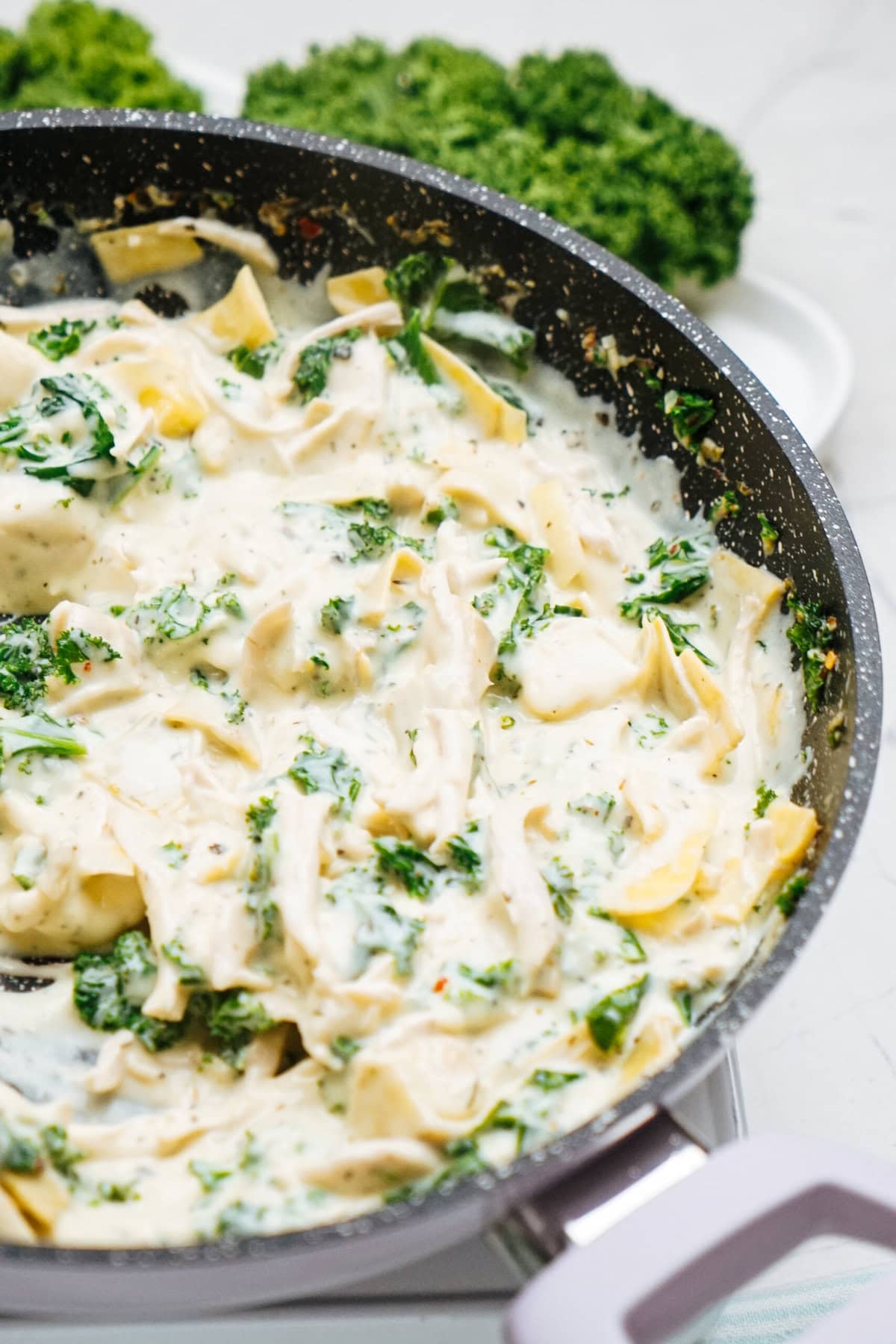 A pan of creamy chicken pasta with spinach and herbs, surrounded by fresh broccoli and kale on a white surface.