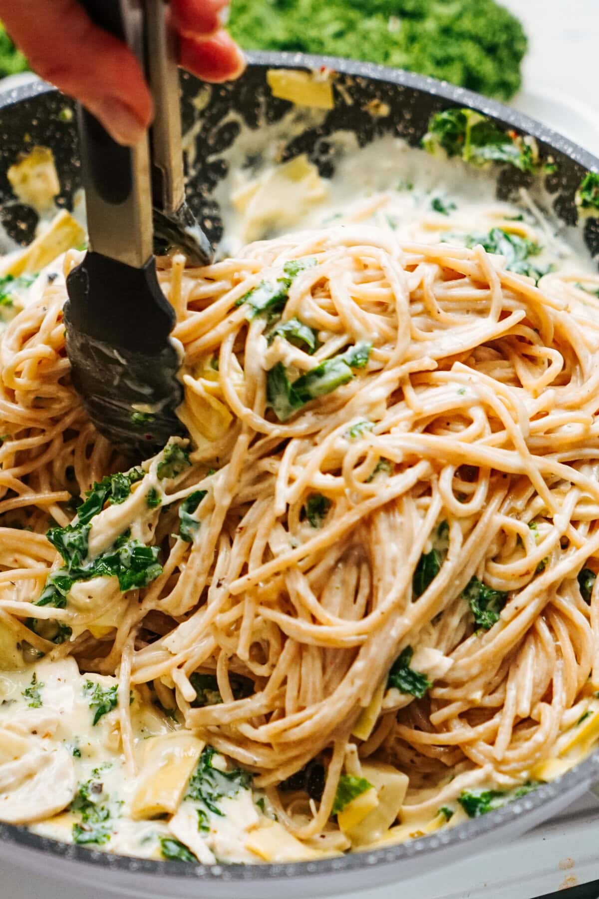 A hand holding tongs stirs a pan of creamy chicken pasta mixed with spinach and artichokes.