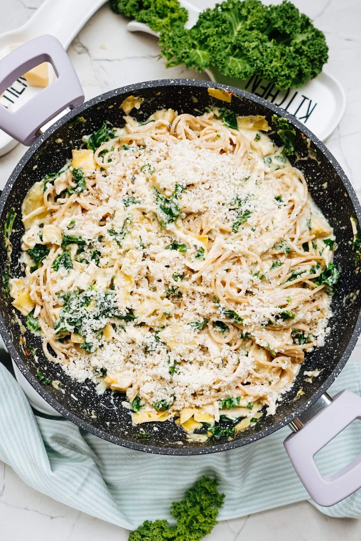 A skillet filled with creamy chicken kale pasta, topped with grated cheese and mixed with spinach and artichokes. Fresh kale is placed nearby on a striped cloth.