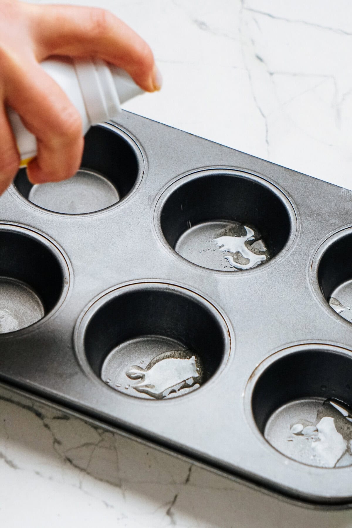 A hand sprays non-stick cooking spray into a muffin tin on a marble surface.