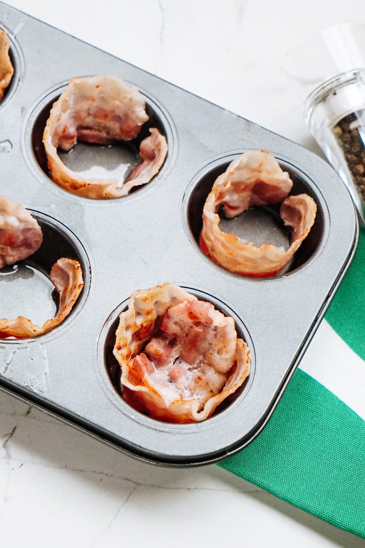 Muffin tin with bacon strips forming cups, placed on a countertop with a striped green and white towel and a pepper grinder beside it.