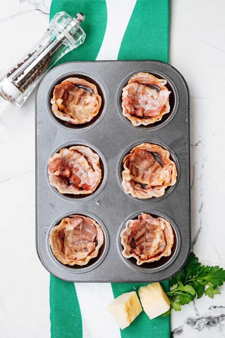 Muffin tin with bacon-wrapped cups on a green and white cloth, next to a pepper grinder, cheese cubes, and parsley on a marble surface.