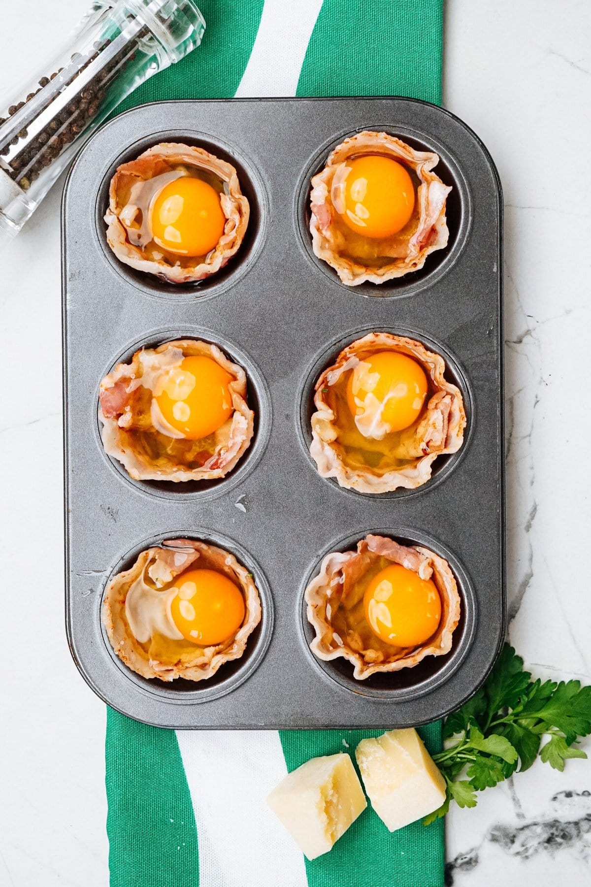 Muffin tin with six uncooked bacon and egg cups, placed on a green and white cloth. A pepper grinder and a cheese wedge are beside the tin.