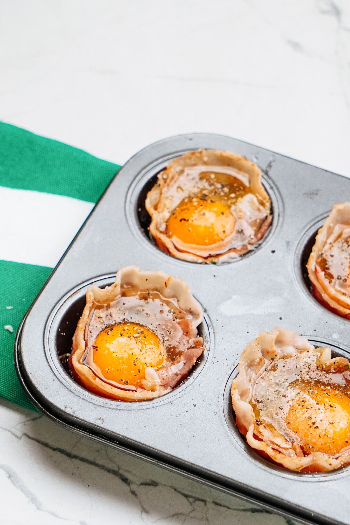 Muffin tin with bacon and egg cups, placed on a marble countertop. A green and white striped cloth is partially visible.