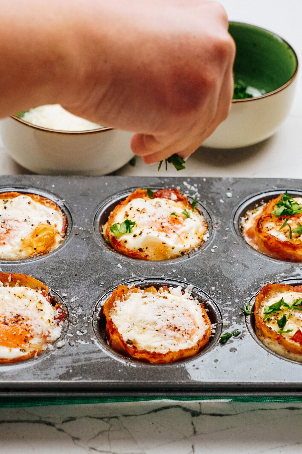 A hand sprinkles herbs over baked egg cups in a muffin tin. Two bowls in the background hold herbs and grated cheese.