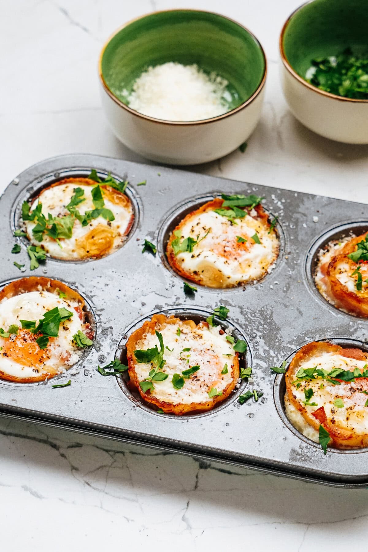 Muffin tin with six baked egg cups topped with herbs. Two small bowls containing grated cheese and chopped greens in the background.