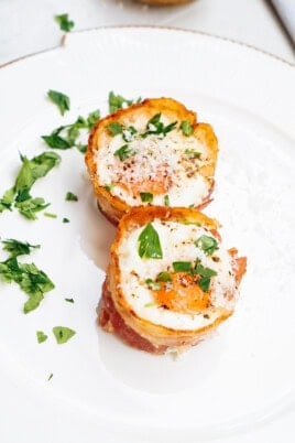 Two bacon-wrapped egg cups topped with chopped parsley and black pepper on a white plate.