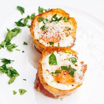 Two bacon-wrapped egg cups topped with chopped parsley and black pepper on a white plate.