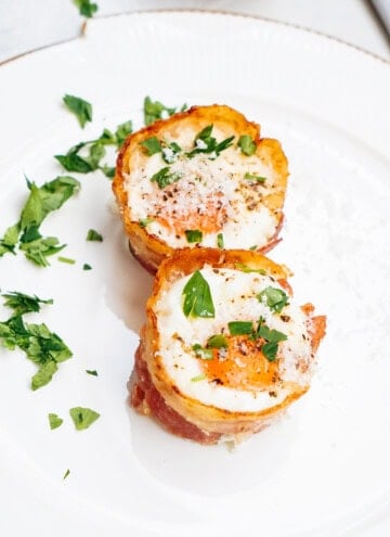 Two bacon-wrapped egg cups topped with chopped parsley and black pepper on a white plate.