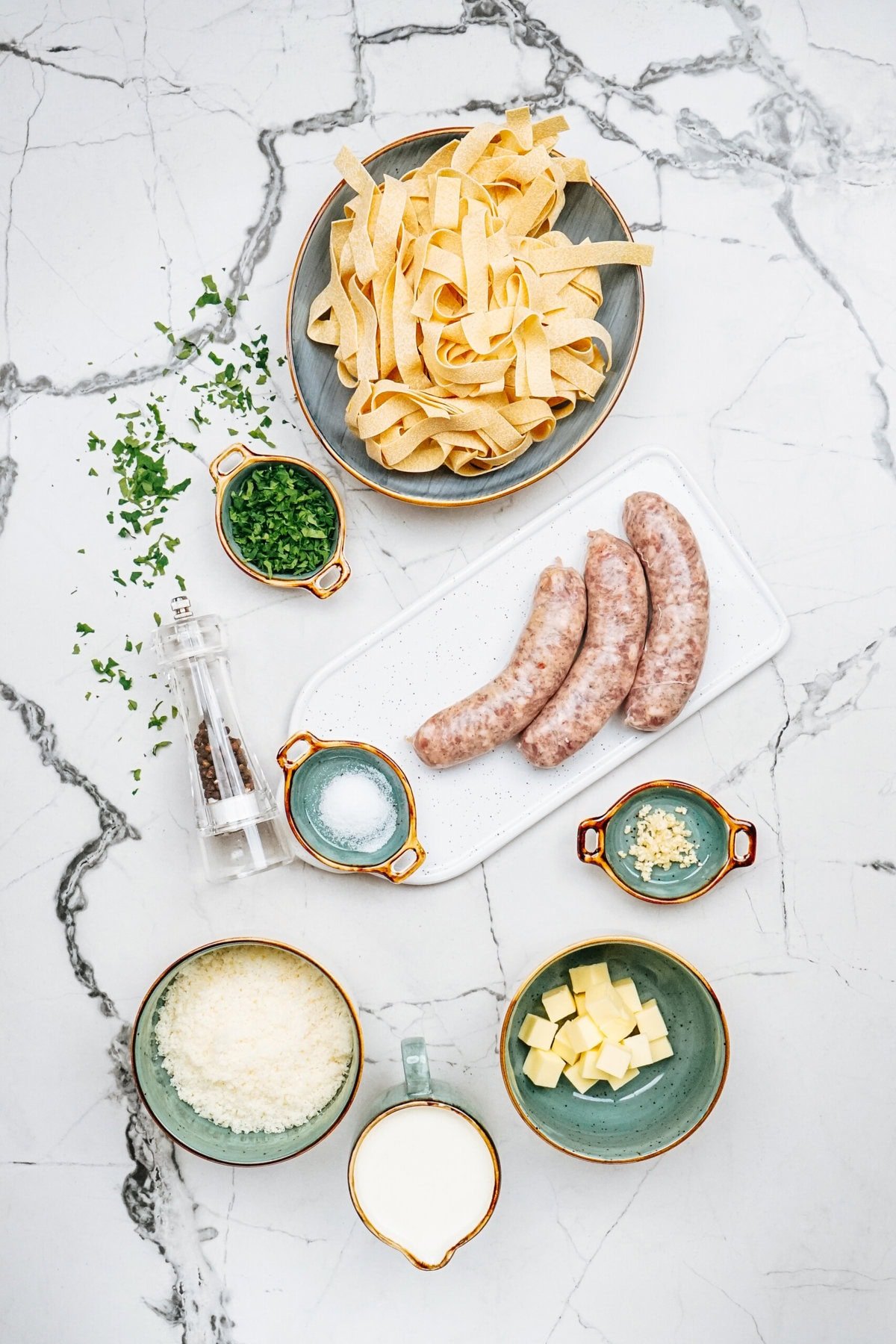 Flat lay of pasta ingredients showcasing the essentials for a delicious Sausage Alfredo: uncooked sausage, fettuccine, chopped herbs, salt, pepper, minced garlic, diced butter, milk, and grated cheese spread across a marble surface.