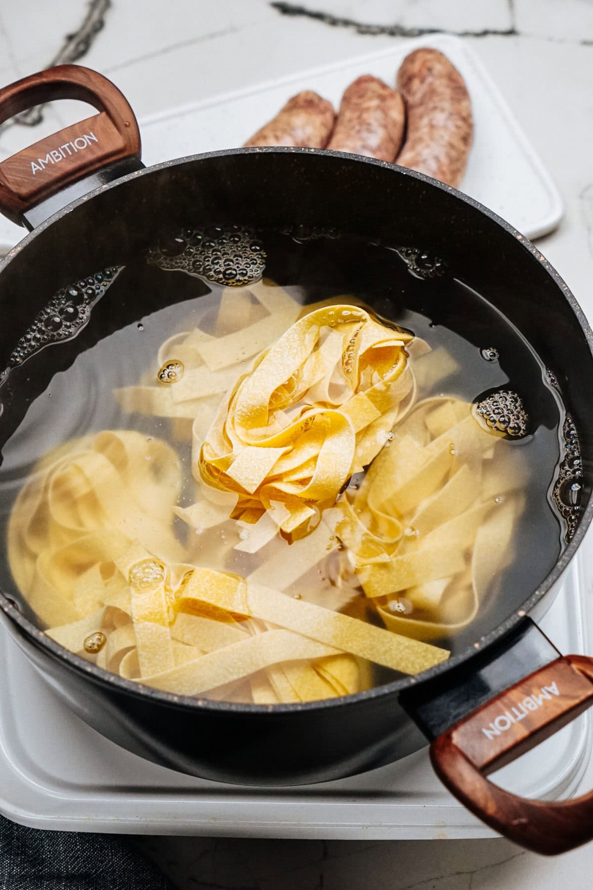 A pot of boiling water holds uncooked pasta ribbons, ready to transform into a delicious Sausage Alfredo. Nearby, raw sausages rest on a plate, waiting to add their savory richness to the dish.