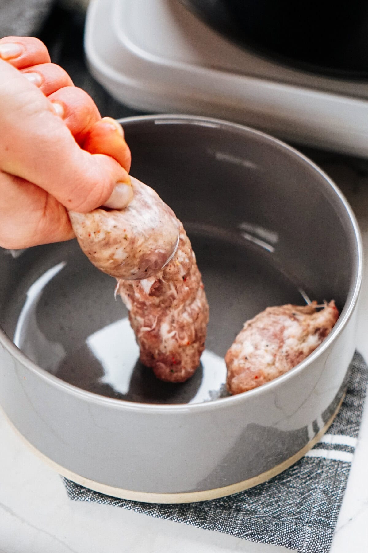 A hand places a raw sausage into a gray bowl on a kitchen counter, preparing for a delicious Sausage Alfredo.