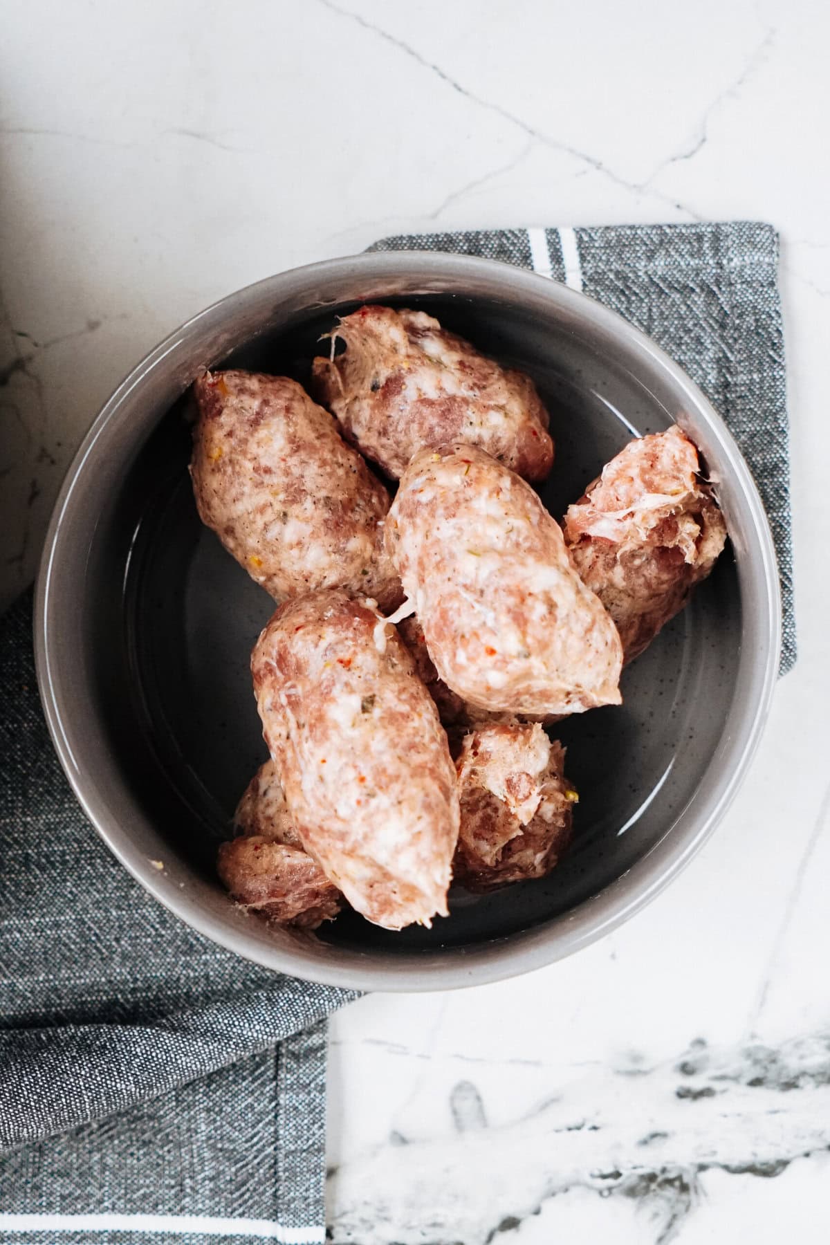 Raw meatballs rest in a gray bowl on a textured gray cloth and marble surface, hinting at the rich flavors of Sausage Alfredo yet to come. .