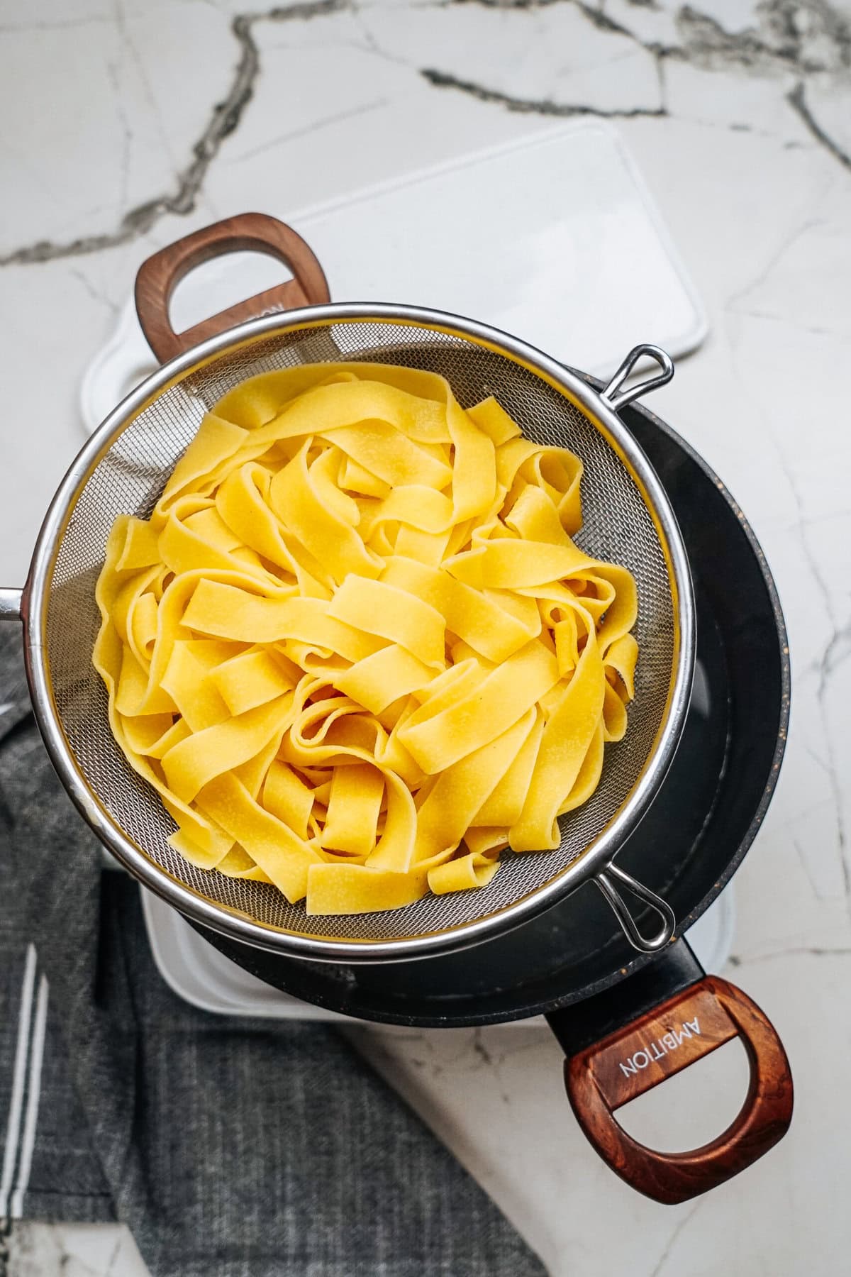 Yellow pasta ribbons in a metal strainer, hinting at a delicious Sausage Alfredo under preparation, sit invitingly over a dark pot on a marble countertop.