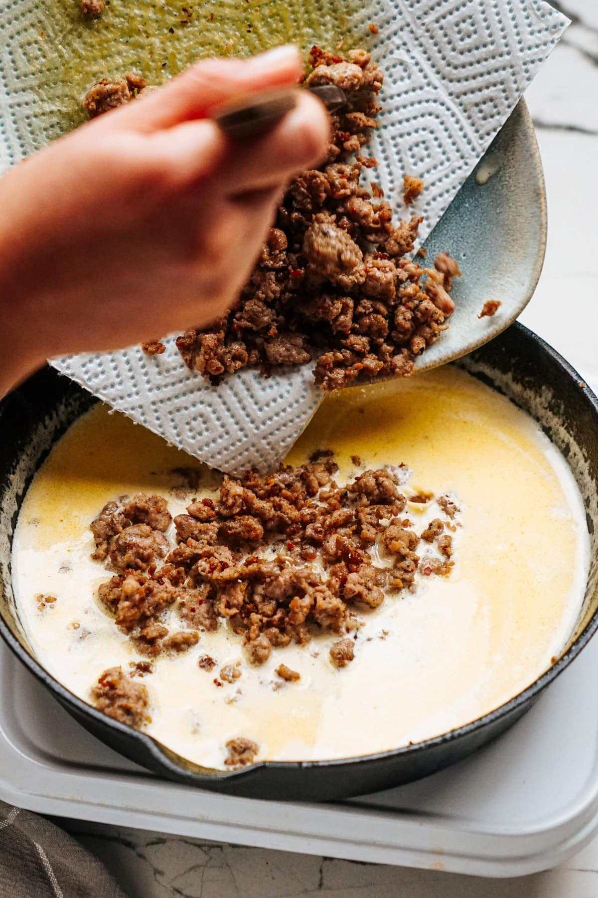 Sausage Alfredo ground meat is being added to a skillet containing a creamy mixture.