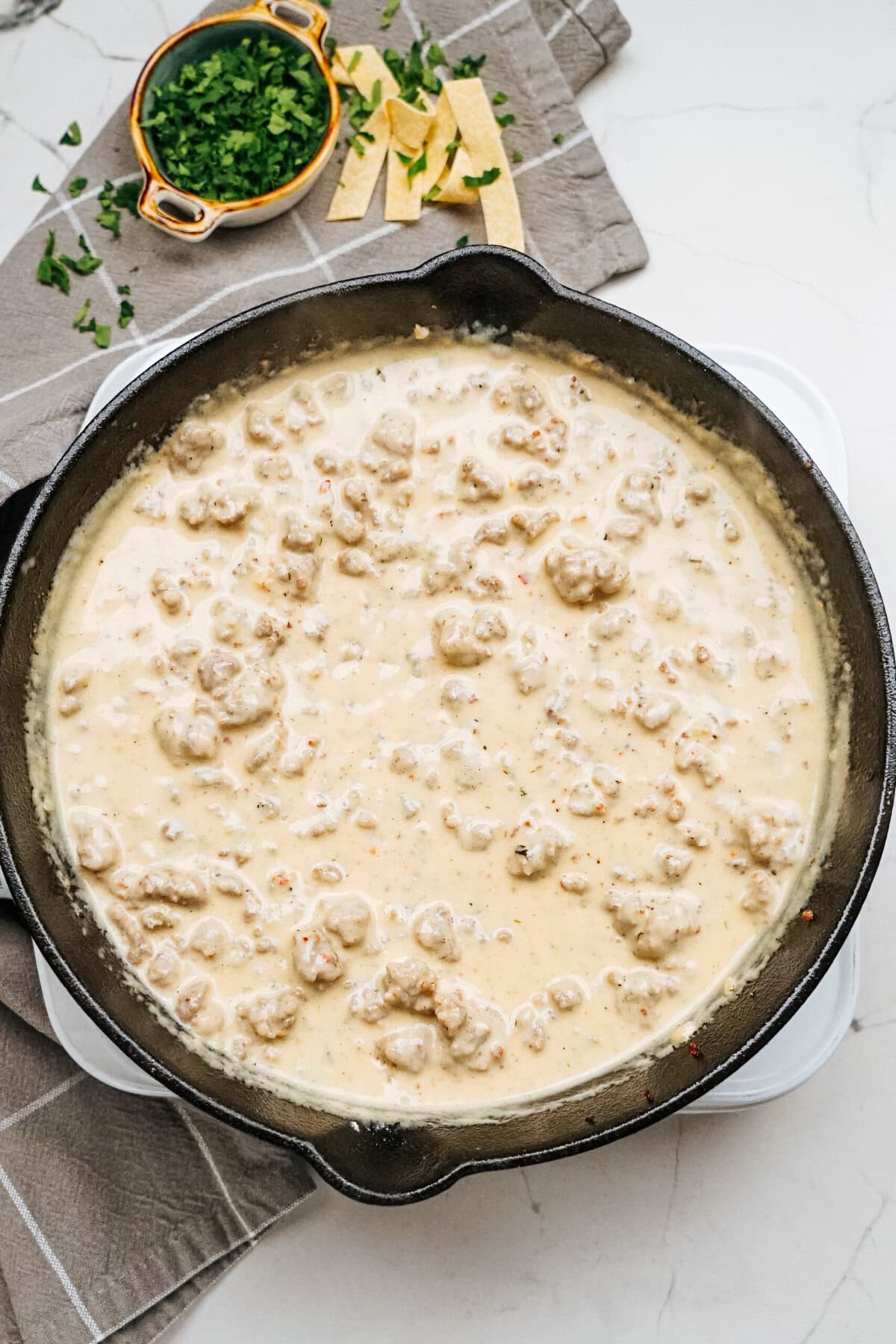 A skillet filled with creamy sausage gravy, reminiscent of a Sausage Alfredo, sits on the table, adorned with chopped parsley and sliced tortilla strips nearby.