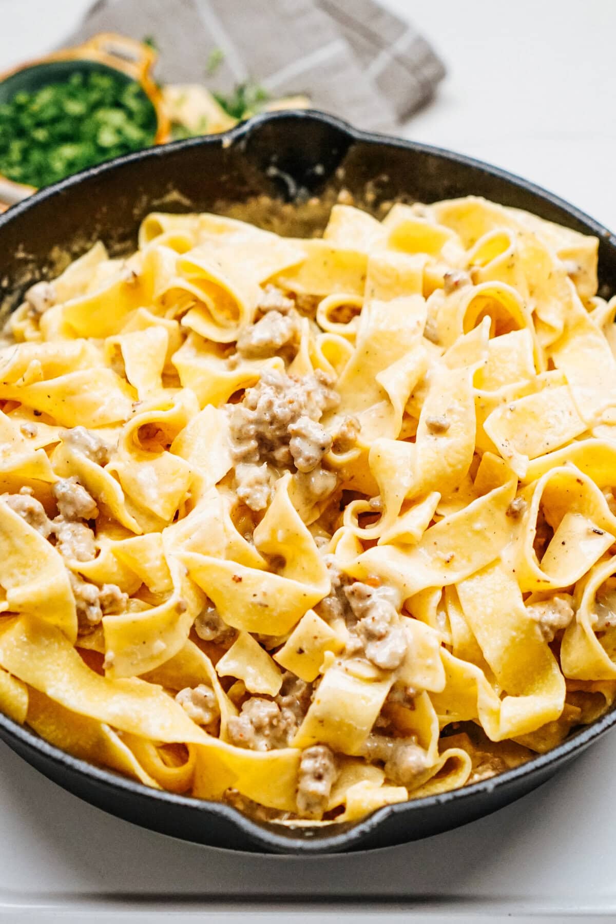 A skillet filled with wide pasta noodles, cooked creamy sauce, and savory ground sausage. A small bowl of chopped herbs is in the background, adding a fresh touch to the indulgent Sausage Alfredo dish.