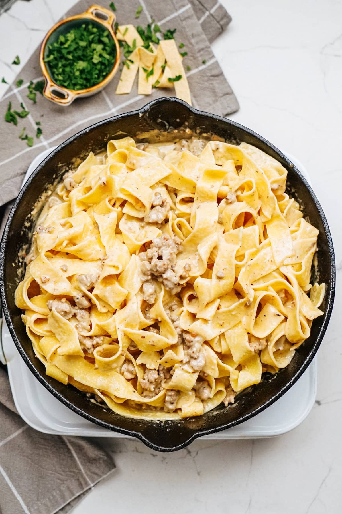 A skillet filled with wide, flat noodles and pieces of sausage in a creamy Alfredo sauce. Chopped herbs are on a small plate beside the skillet, placed on a cloth napkin.