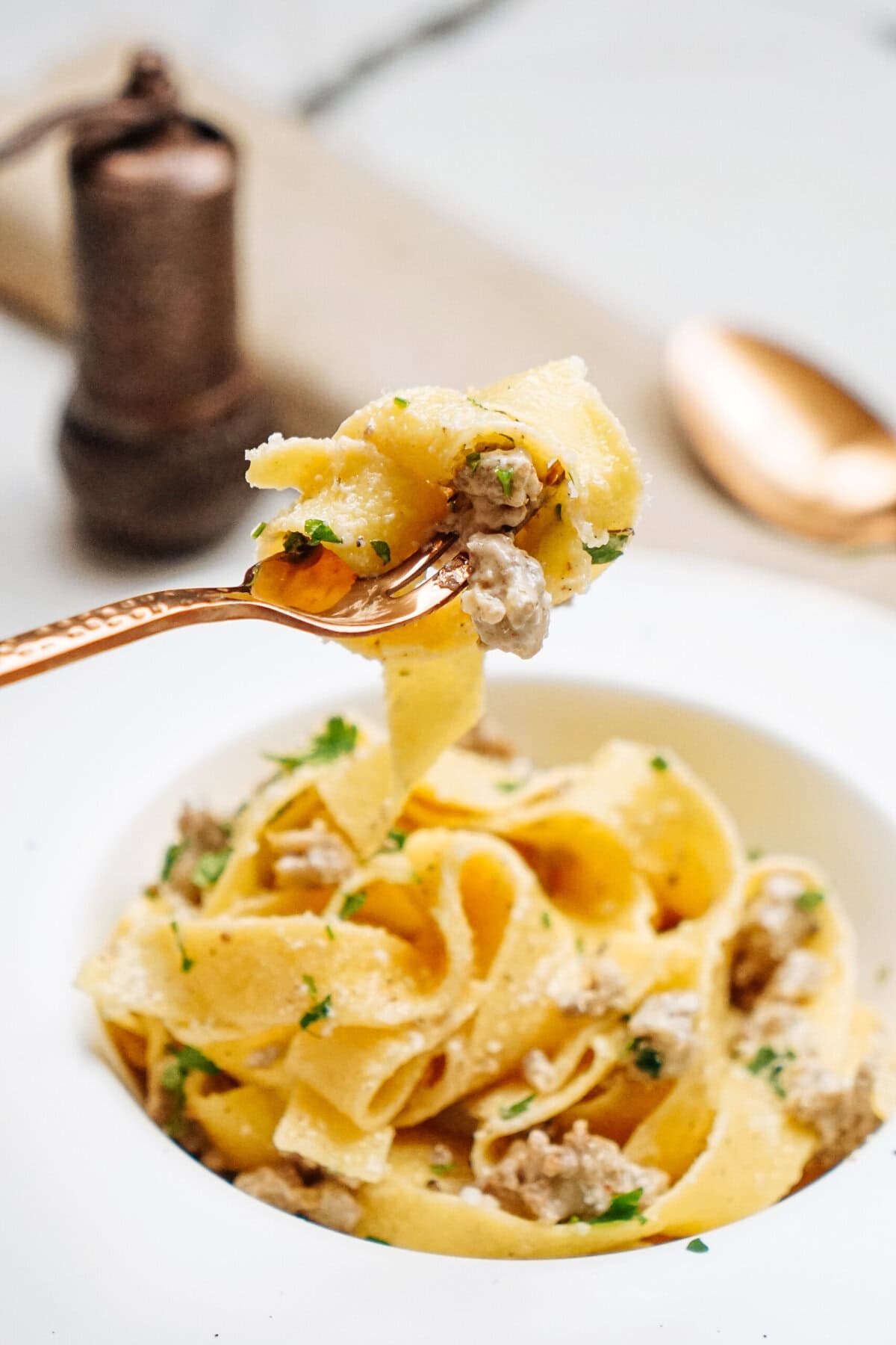 A fork holds a portion of creamy sausage Alfredo with herbs and meat, above a white bowl filled with the same dish. A pepper grinder and a spoon are blurred in the background.