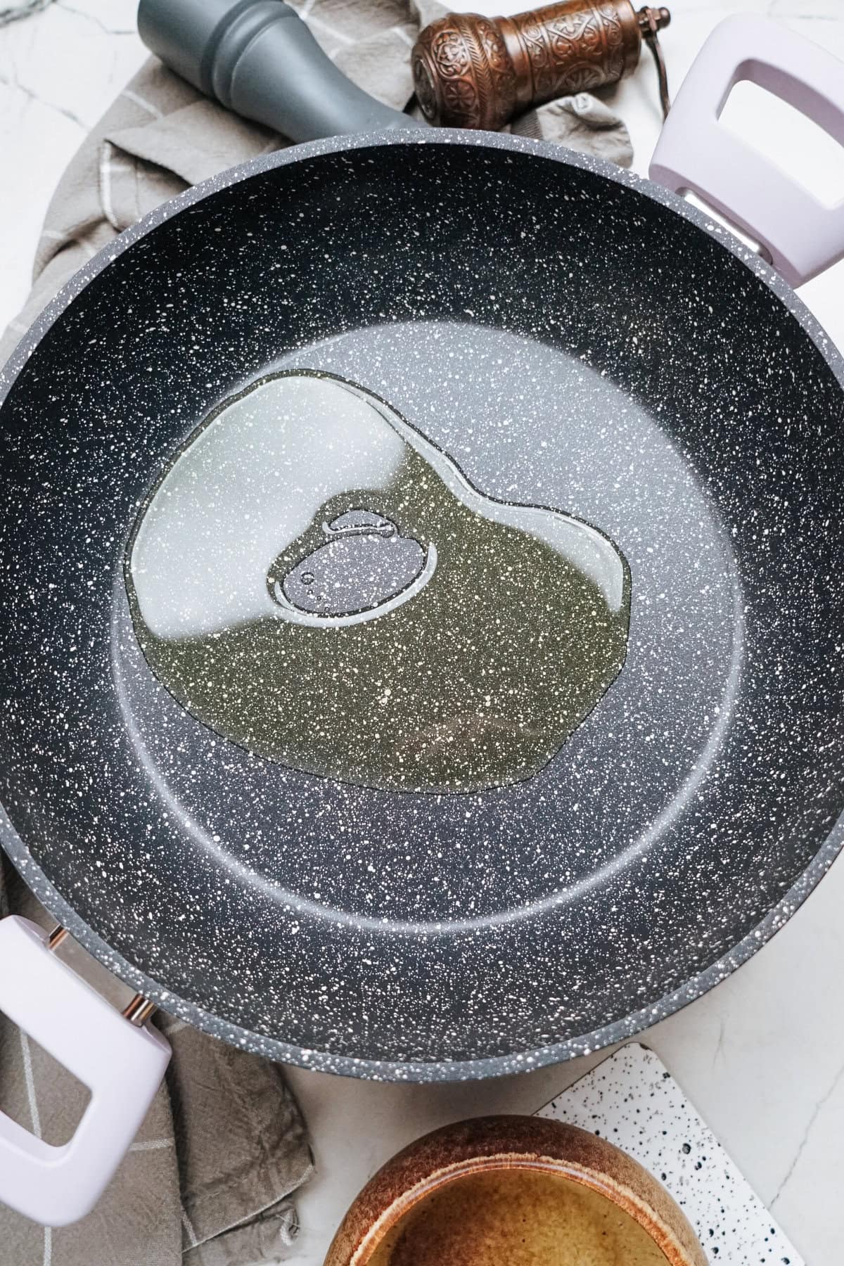 A black frying pan with oil heating inside, ready for a stir fry, surrounded by a brown bowl, pepper grinder, and cloth on a white marble surface.