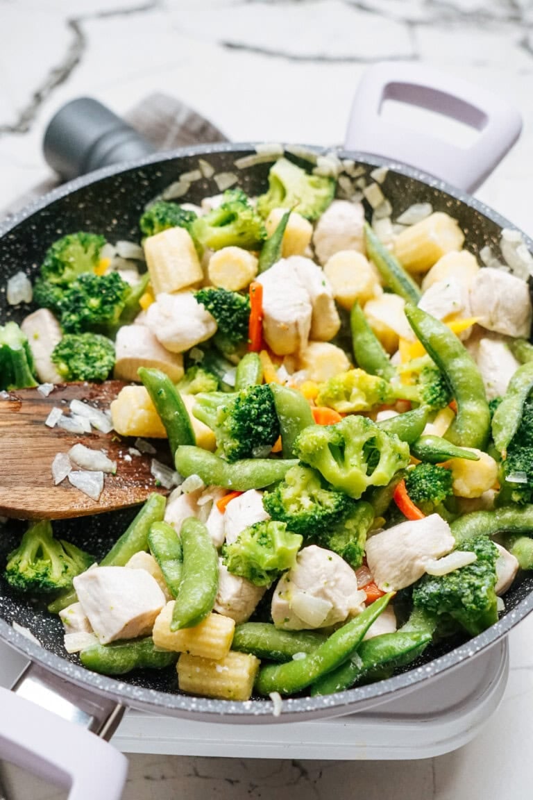 A pan sizzles with a vibrant stir fry of broccoli, snap peas, baby corn, bell peppers, and chunks of chicken, all expertly tossed with a wooden spatula.