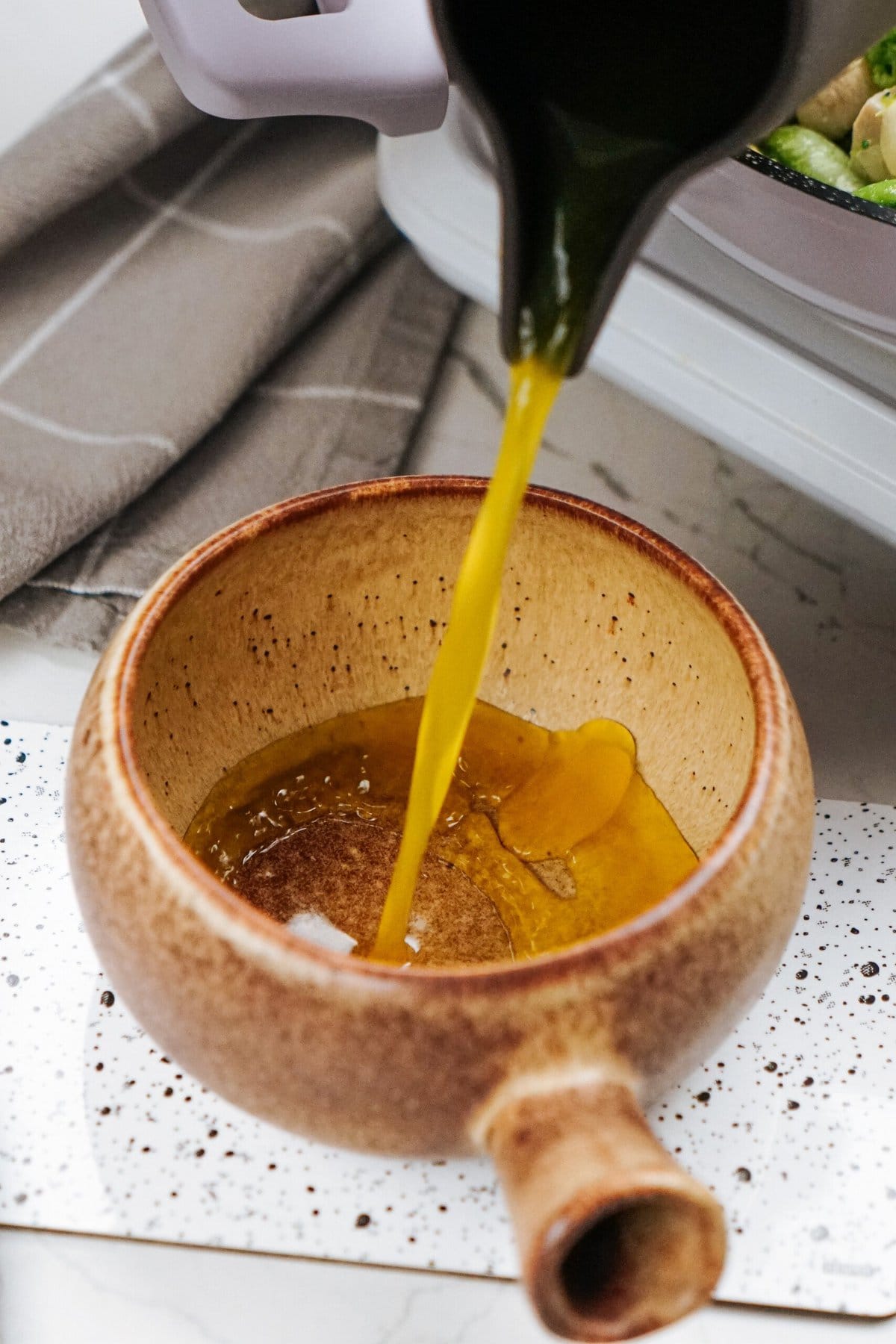 Oil being poured from a black container into a brown ceramic pot on a speckled surface, ready to enhance a flavorful stir fry, with a gray cloth in the background.