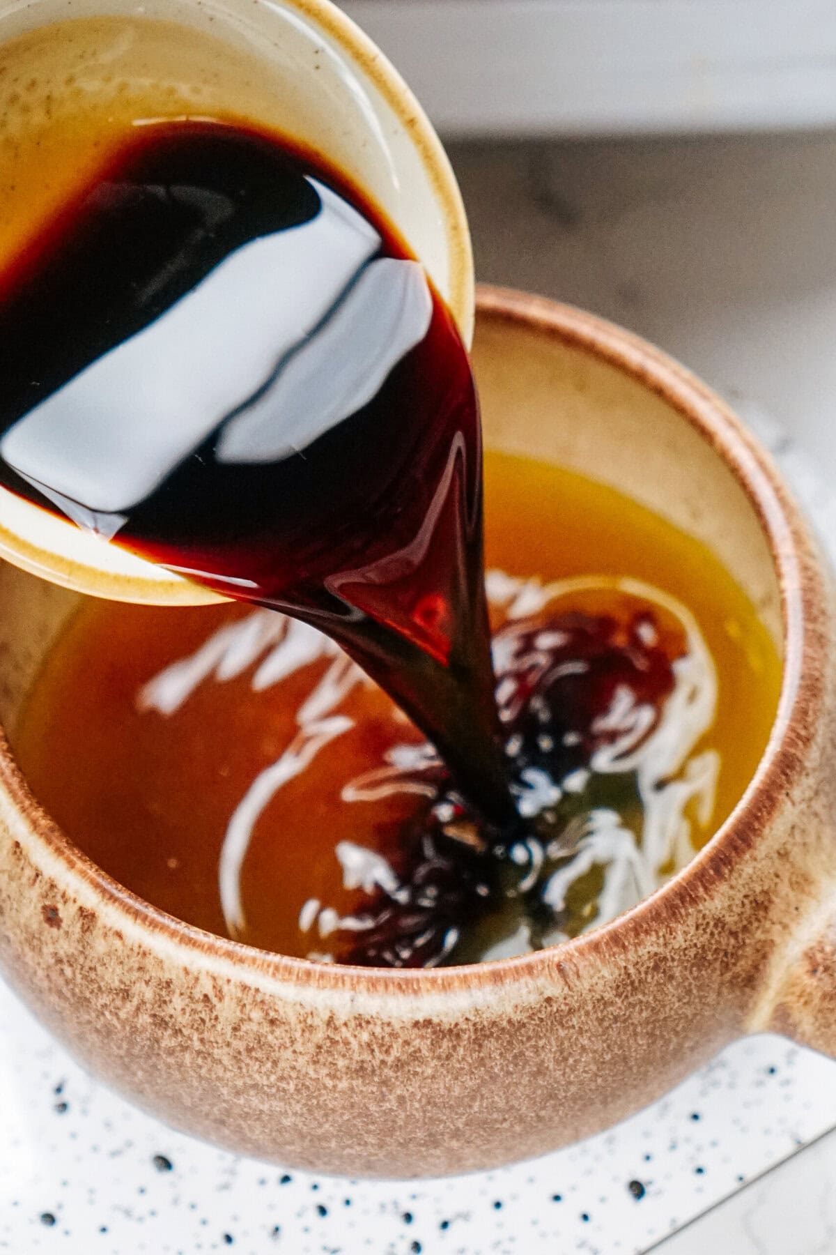 Dark liquid is being poured from a cup into a brown ceramic bowl, reminiscent of the rich sauces often used in a stir fry dish.