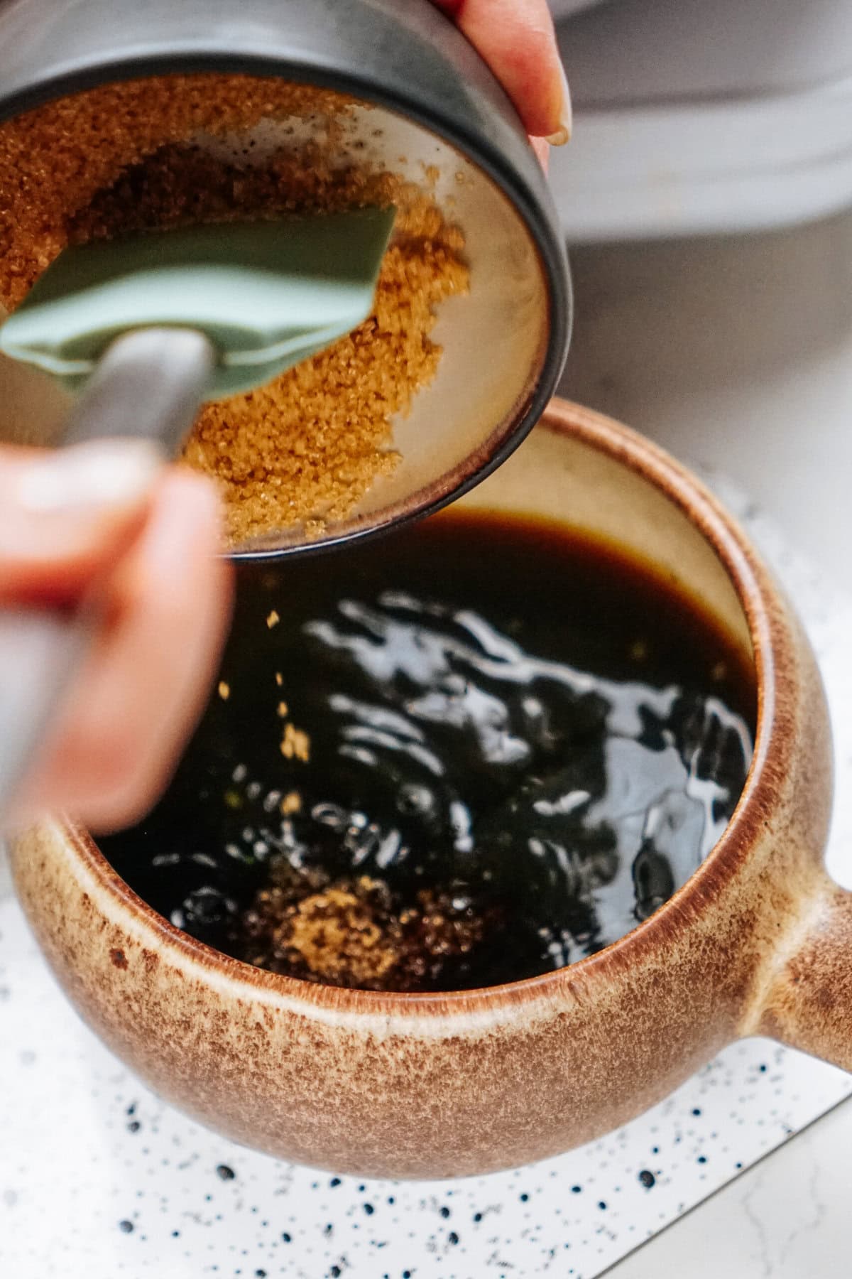 A hand uses a green spatula to pour brown sugar into a brown bowl filled with dark liquid on a speckled surface, as if preparing the perfect sauce for an irresistible stir fry.