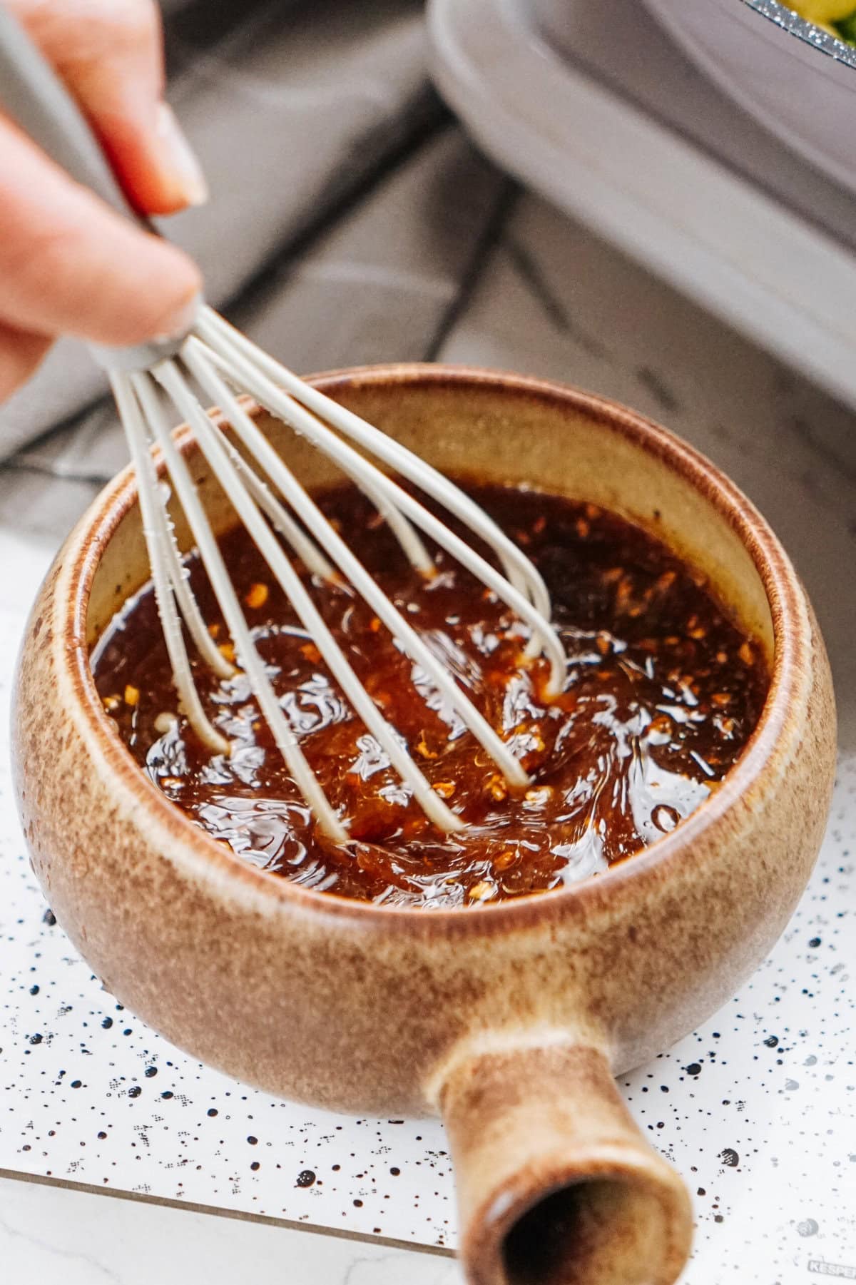 A small ceramic bowl holds a dark stir fry sauce, being whisked by hand with a metal whisk. The sauce, teeming with visible herbs or spices, rests invitingly on a speckled surface.