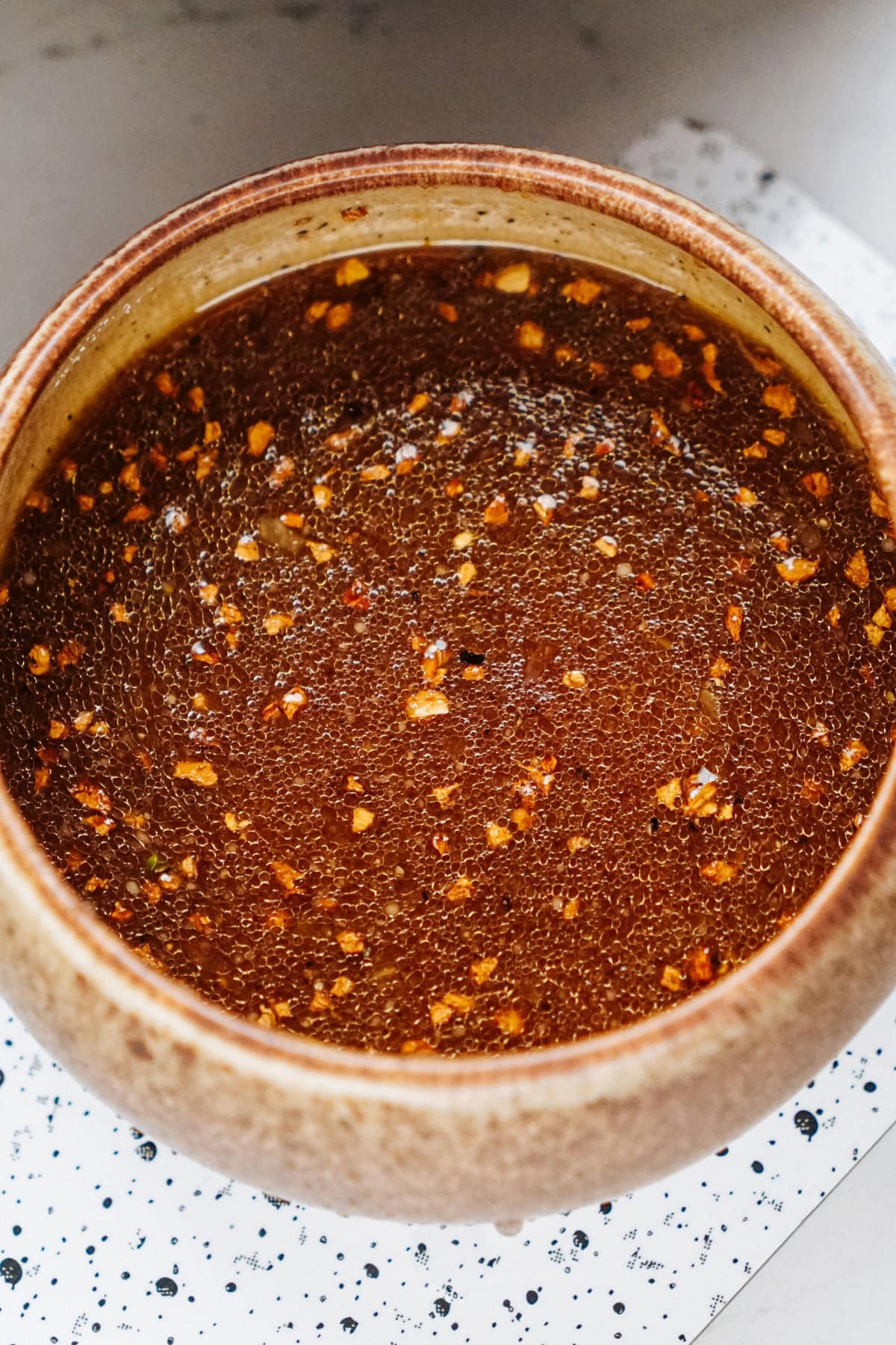 A bowl filled with a savory brown stir fry sauce, peppered with red flakes and small bits of other spices, sits on a white surface dotted with black specks.