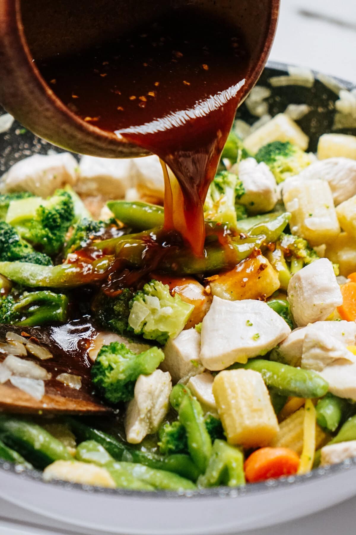 A rich sauce cascades over a sizzling stir-fry, filled with vibrant broccoli, tender baby corn, crisp carrots, and juicy chicken pieces in the pan.