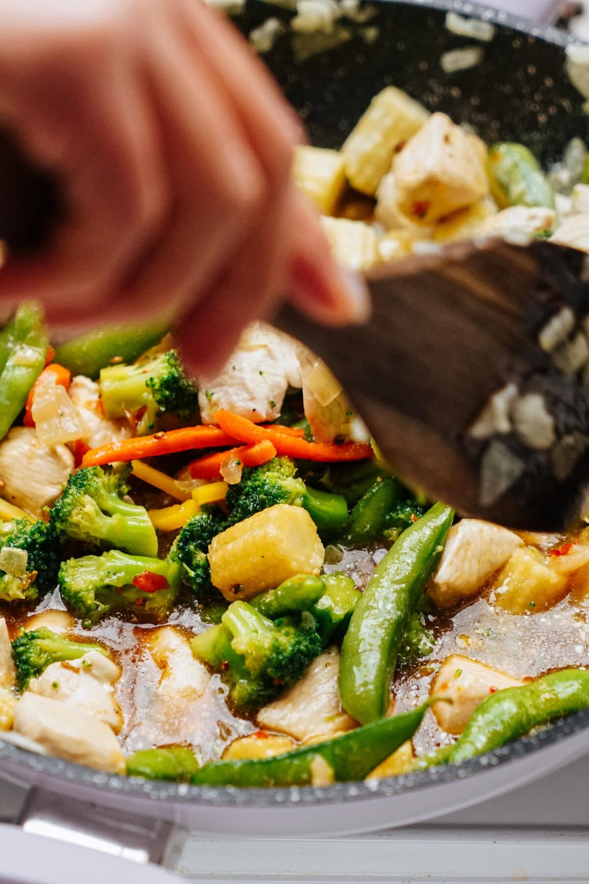 A person expertly stirs a vibrant stir-fry in the pan, combining colorful vegetables, tender chicken, and juicy pineapple using a wooden spatula.