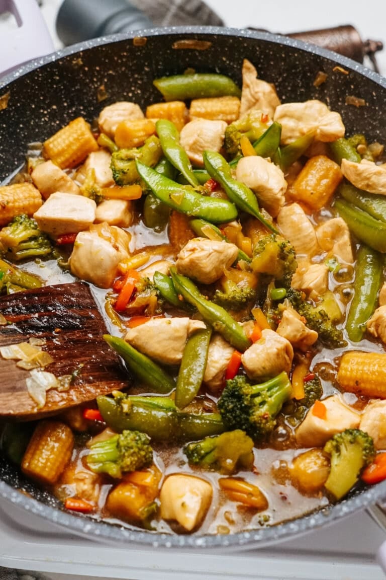 A skillet filled with a vibrant stir fry featuring chicken pieces, broccoli, sugar snap peas, baby corn, and diced red peppers in a savory sauce. A wooden spatula rests on the side of the pan.