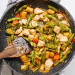 A vibrant stir fry featuring chicken, broccoli, snap peas, baby corn, and carrots dances in a sizzling skillet, with a trusty wooden spatula at the ready.